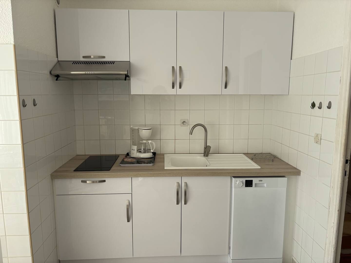 Eine kompakte Küche mit weißen Schränken, einer Edelstahl-Dunstabzugshaube, einer Kaffeemaschine, einem Spülbecken und einem Geschirrspüler.