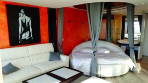 Moderni hotellihuone, jossa on kaareva sänky, valkoinen sohva, lasinen sohvapöytä ja suuri taulu seinällä.