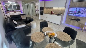 A modern living room with a sofa, chairs, and a dining table with a fruit bowl. There is a kitchen area with cabinets and a TV displaying a show. - HOLIDAYS - Studio Héliopolis C 66 - Luxury - 2 people (AGDE)
