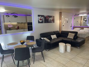 Ein modernes Wohnzimmer mit einem schwarzen Ecksofa, einem Glastisch mit gewebten Platzsets und einer Küche im Hintergrund.