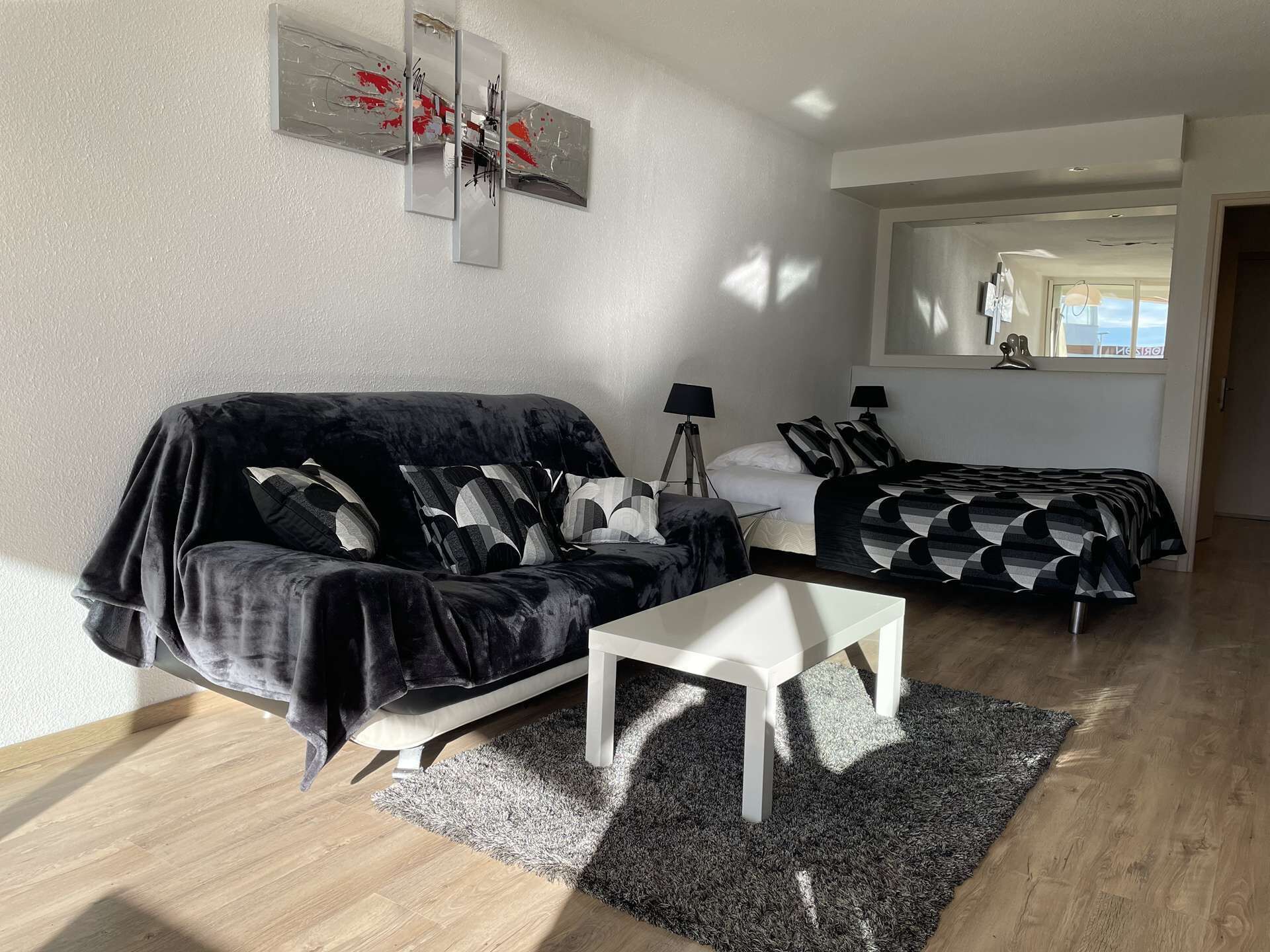 A modern living room with a black sofa, a white coffee table, and a bed with patterned bedding.