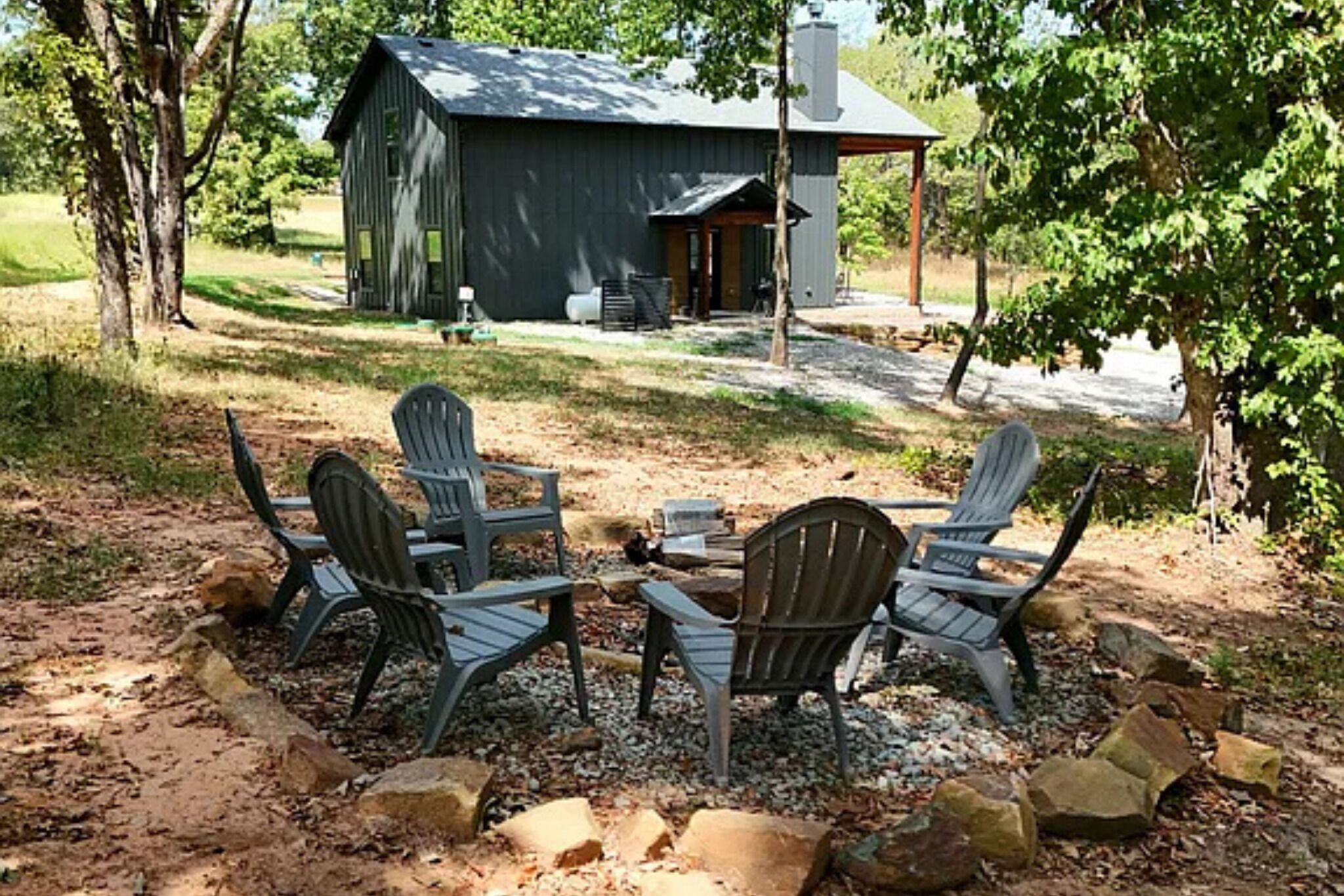 A rustic outdoor seating area with Adirondack chairs around a small fire pit, a wooden deck, and a large barn-like structure in the background.