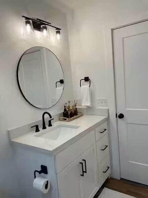Une salle de bain avec un lavabo blanc, un robinet noir et un miroir rond. Trois lampes sont placées au-dessus du miroir, et on peut voir un porte-serviettes.