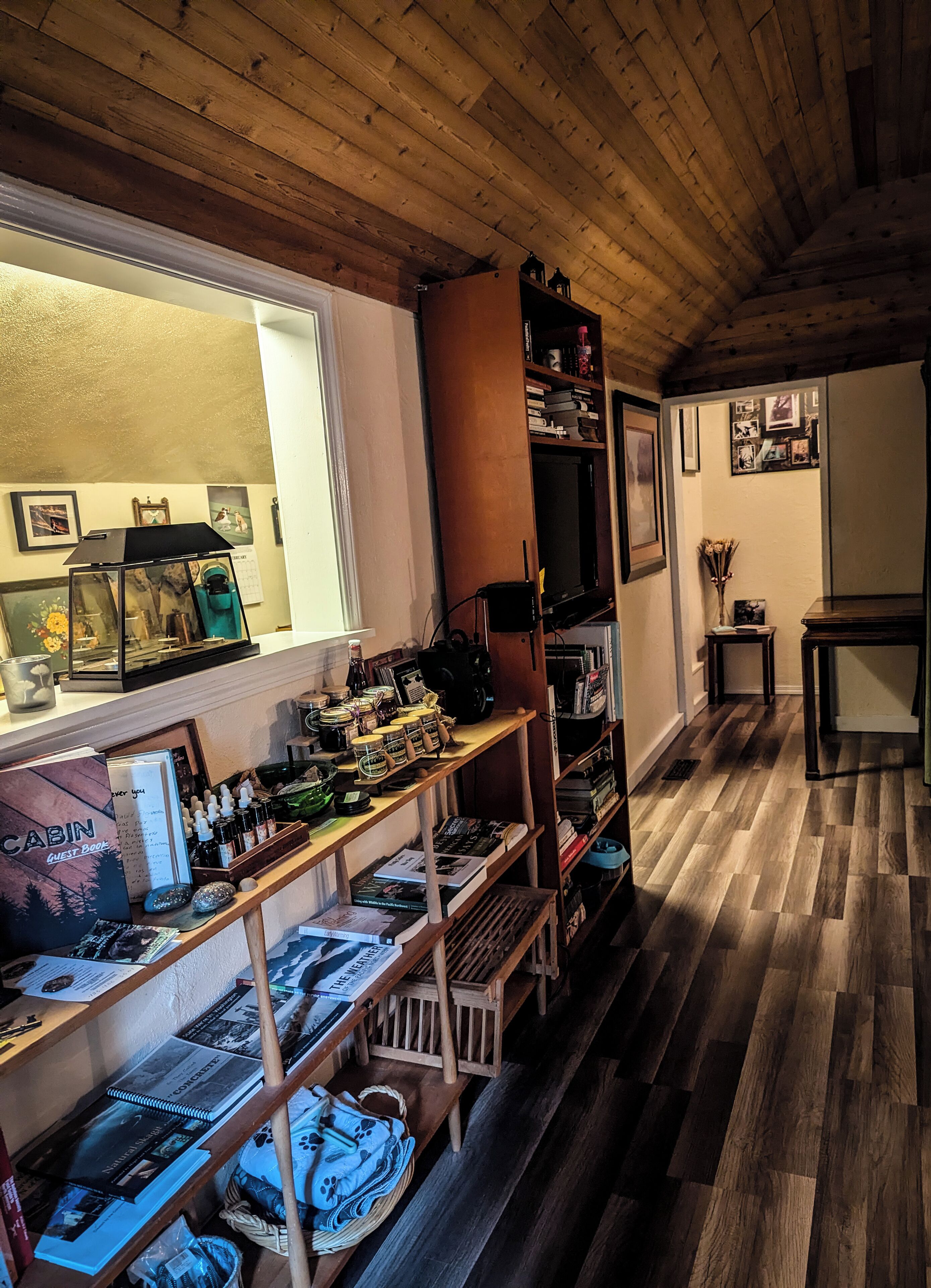 A cozy cabin interior with wooden floors, a bookshelf, a desk with various items, and a window with framed pictures.