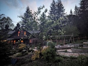 A rustic cabin with a balcony, surrounded by dense forest and a garden with various plants and a wooden fence. - Whimsical and cozy cottage on a homestead in the North Cascades (Concrete)