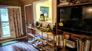 A cozy room with wooden floors, a bookshelf filled with books and games, a television, and a window with curtains. - Whimsical and cozy cottage on a homestead in the North Cascades (Concrete)