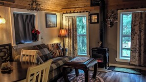 A cozy cabin interior with a wooden ceiling, a dining table with chairs, a sofa, a fireplace, and a window with curtains. - Whimsical and cozy cottage on a homestead in the North Cascades (Concrete)