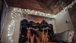 A bedroom with a bed, a mountain-themed tapestry, string lights, and a pillow. - Whimsical and cozy cottage on a homestead in the North Cascades (Concrete)