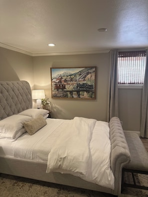 A neatly made bed with a headboard, a bedside table with a lamp, and a painting of a mountainous landscape on the wall.