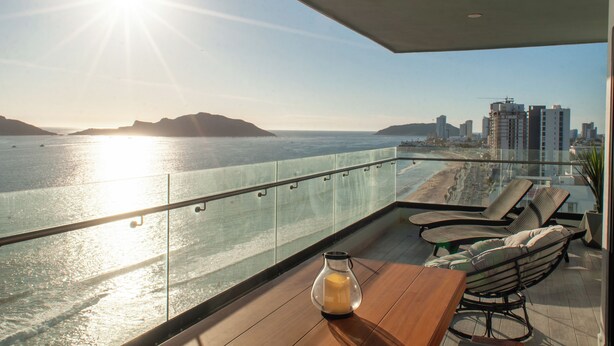 Un balcón con vistas al mar y al paisaje urbano, que incluye una mesa de madera y dos sillones reclinables.