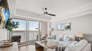 Ein geräumiges Wohnzimmer mit einer großen Couch, einem Couchtisch und einem Deckenventilator. Eine Glastür öffnet sich zu einem Außenbereich mit Blick auf die Landschaft.