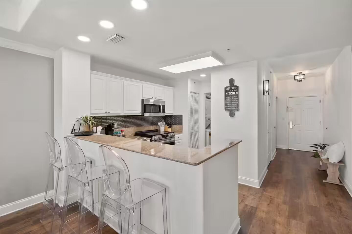 Uma cozinha moderna com armários brancos, uma ilha central com banquetas, micro-ondas e uma placa com a inscrição 'Welcome to the Kitchen'.
