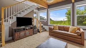 A living room with a staircase, a television, a leather sofa, and large windows. - Castello Del Mare - Ocean views, Hot tub, Dogs OK (Newport)