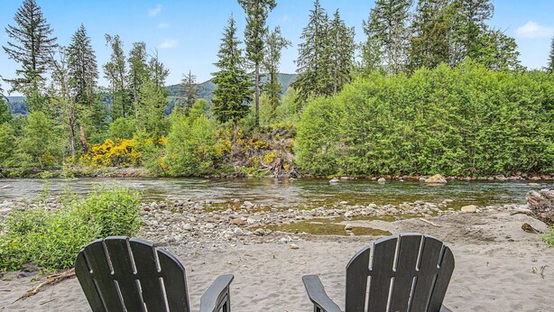 A river with a rocky bed, surrounded by dense green trees and a clear sky. Two black chairs are placed on the sandy shore.