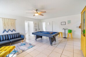 A room with a pool table, a sofa, a ceiling fan, and a green bag on a stool.