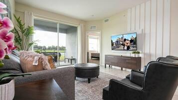 Un salon moderne avec un canapé, un pouf et des fauteuils en cuir noir. Un téléviseur à écran plat est fixé au mur, et une porte-fenêtre coulissante donne accès à un balcon d’où l’on peut admirer la verdure.