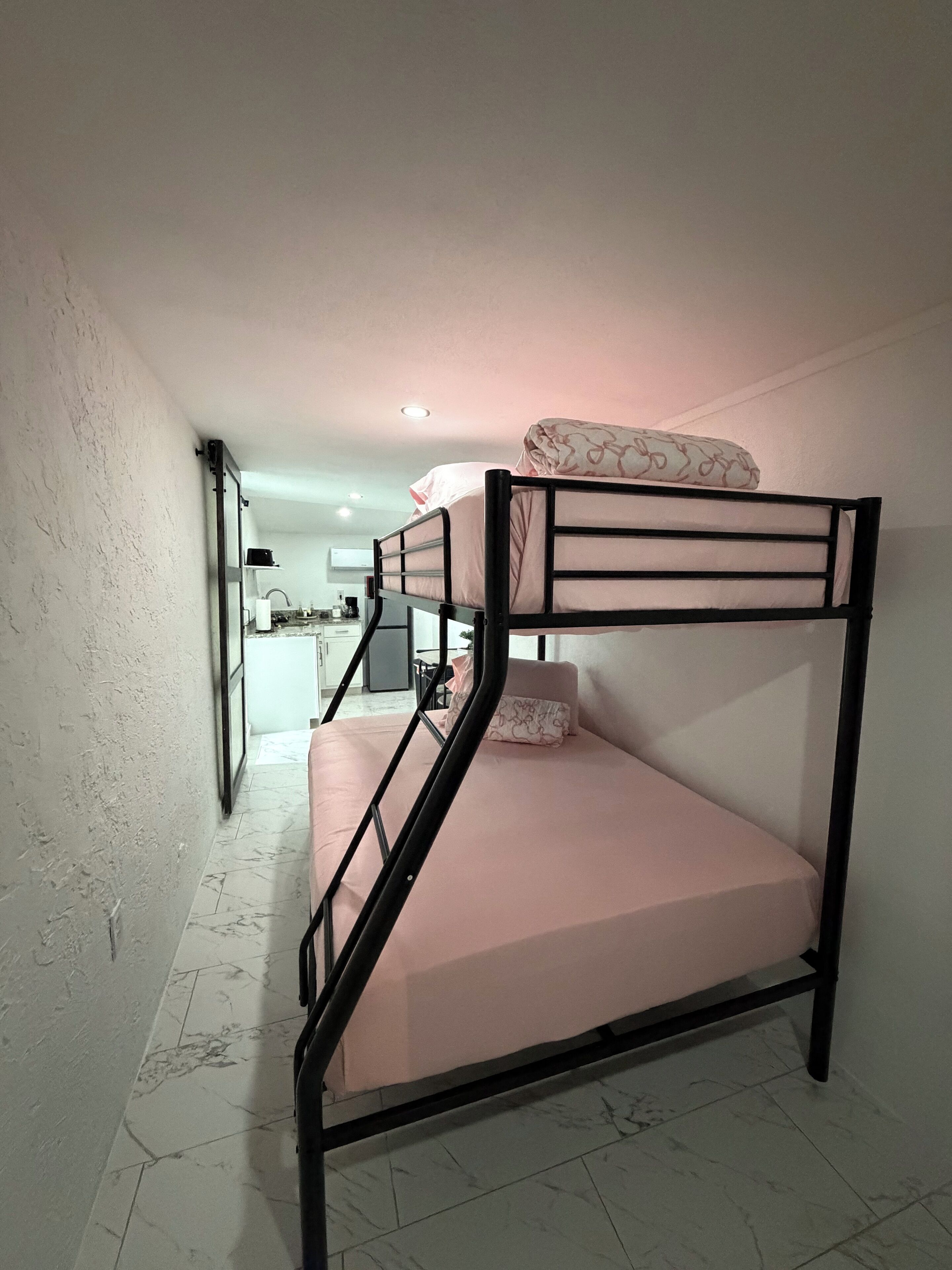 Un lit superposé avec un matelas rose et des oreillers à motifs, un mur blanc et un sol carrelé.