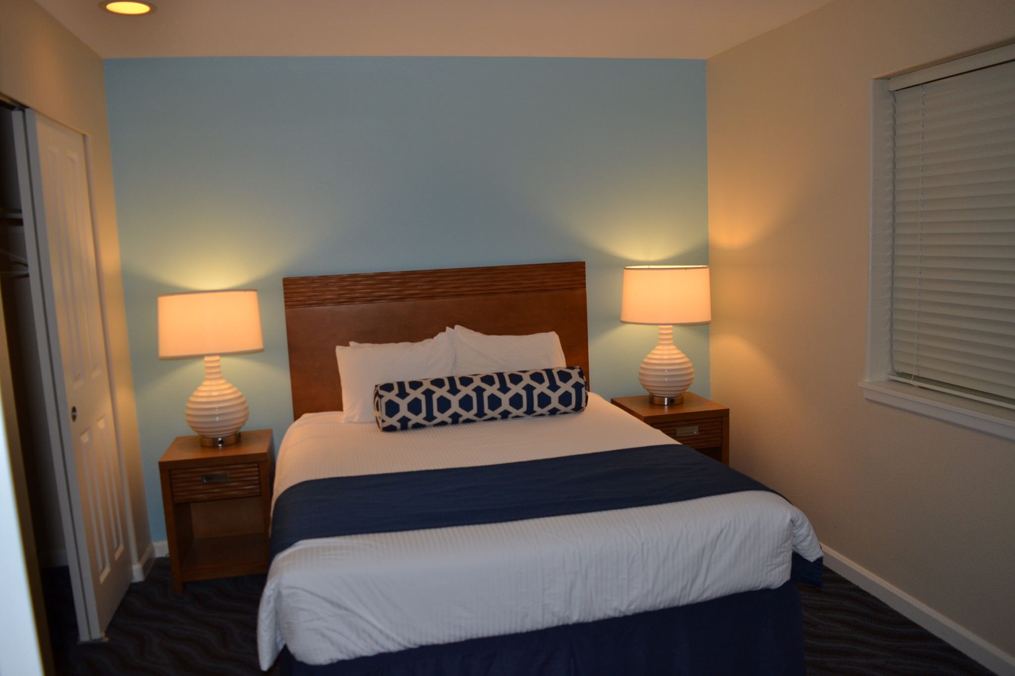 A neatly made bed with a wooden headboard, two bedside lamps, and a window with blinds.