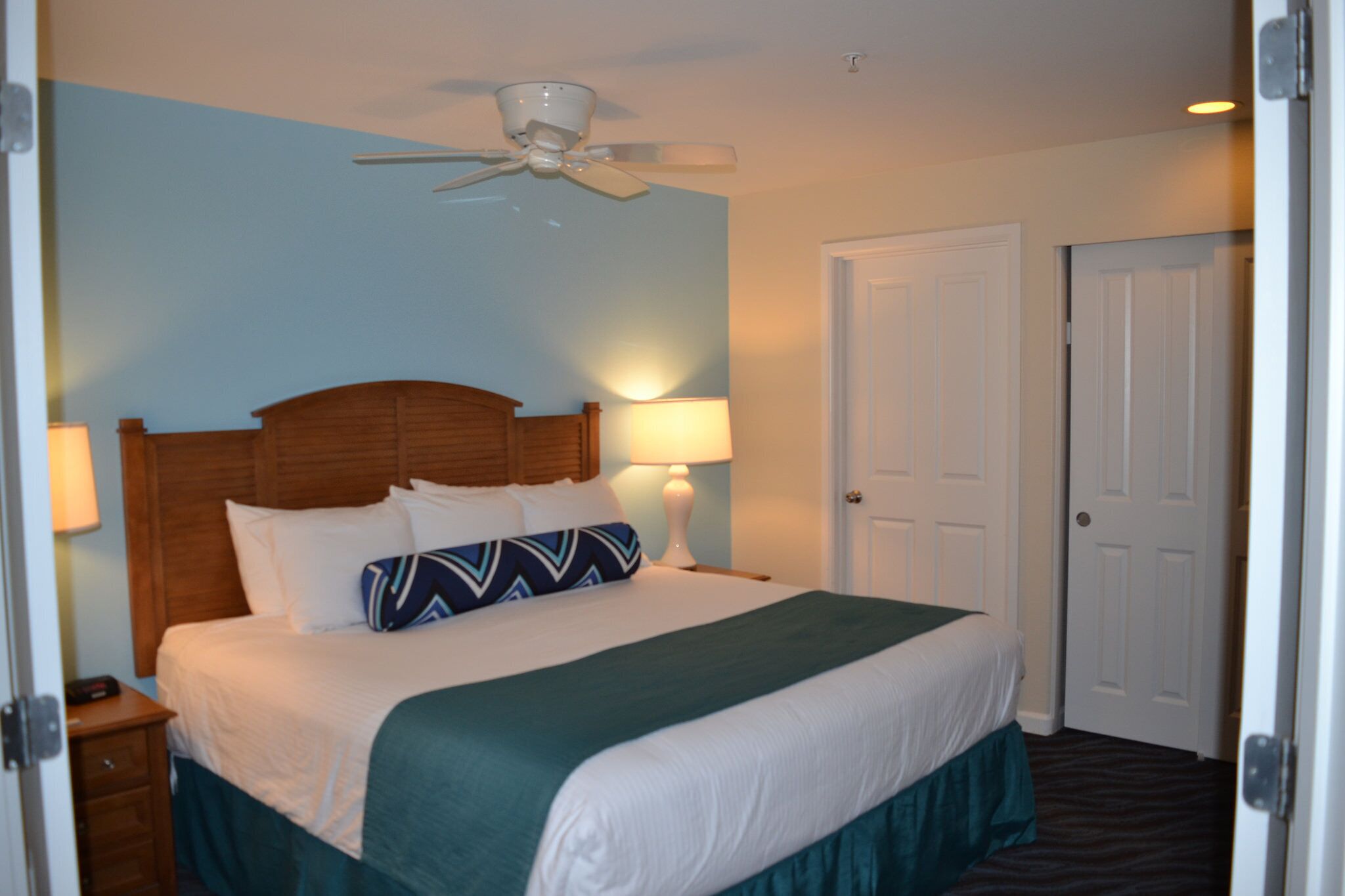 A hotel room with a single bed, a ceiling fan, a nightstand with a lamp, and a door.
