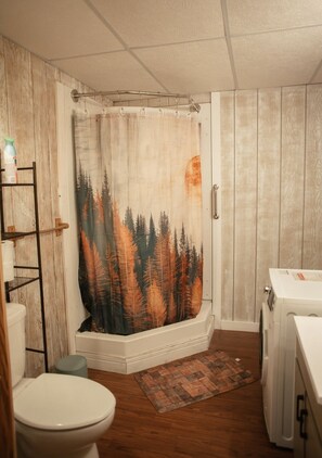 Un baño con cortina de ducha que muestra un paisaje de bosque, un inodoro, una lavadora y piso de madera.