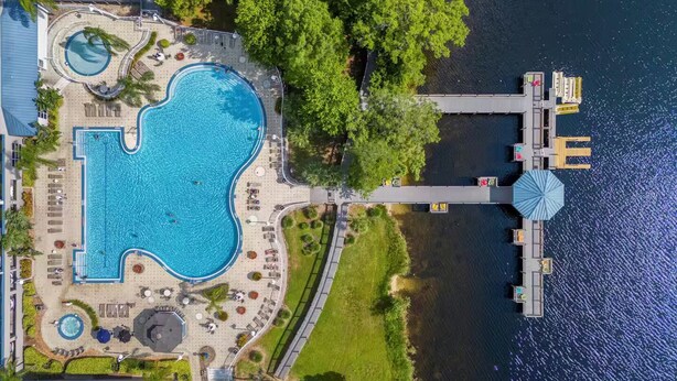 An aerial view of a resort with a large central swimming pool, a dock extending into a body of water, and surrounding greenery. - Trendy Lake Condo-Disney view! (Orlando)