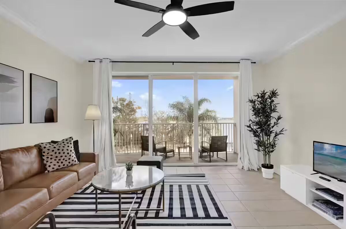 A modern living room with a brown leather sofa, a round coffee table, a TV on a white stand, a ceiling fan, and a balcony with outdoor furniture.