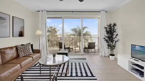 A modern living room with a brown leather sofa, a round coffee table, a TV on a white stand, a ceiling fan, and a balcony with outdoor furniture. - Trendy Lake Condo-Disney view! (Orlando)