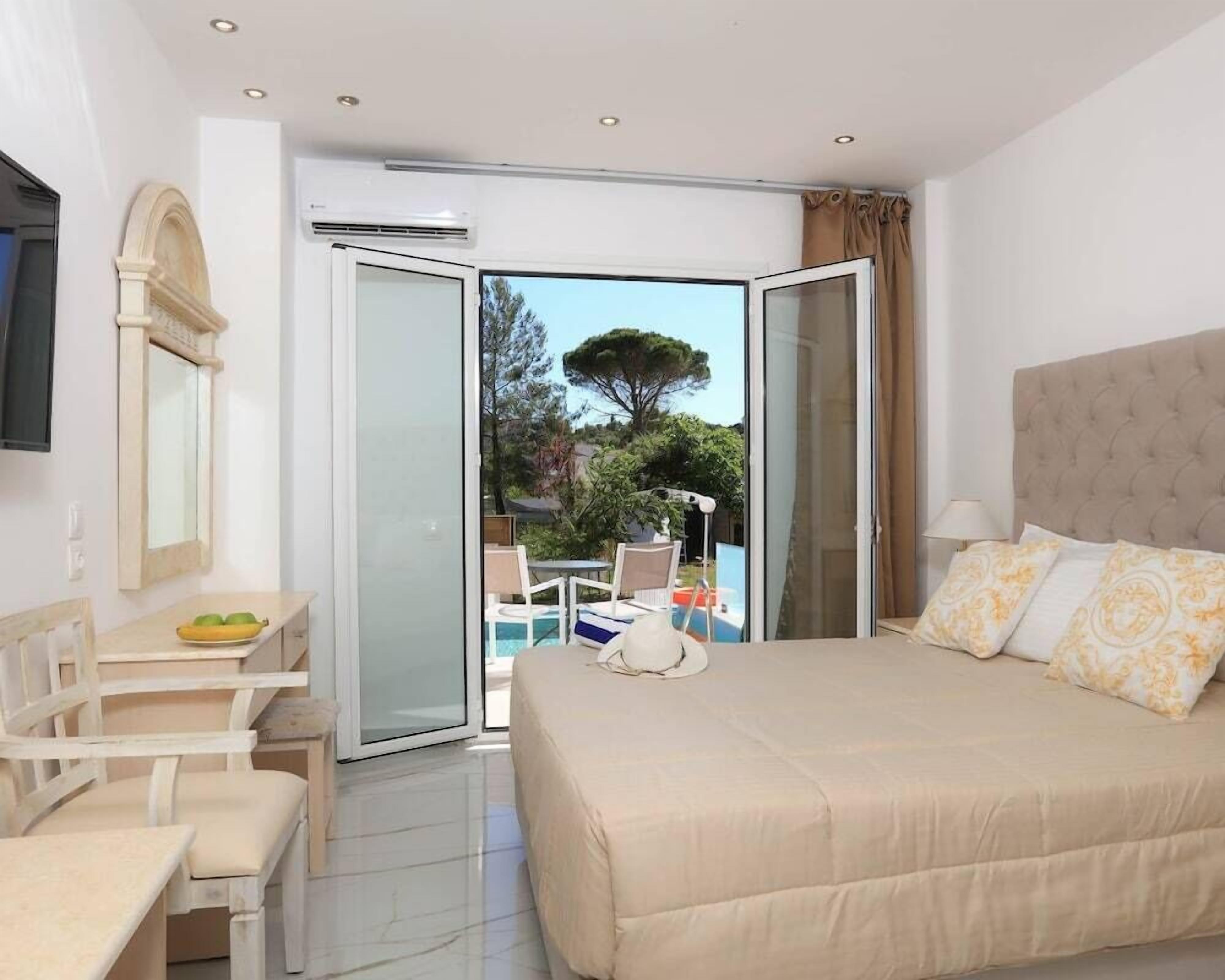 Habitación de hotel con cama, escritorio con sillas, mesa de comedor y vistas a una piscina y árboles.