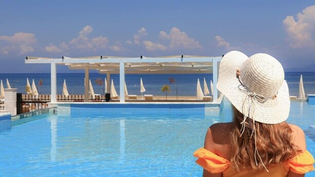 Une vue sur la piscine avec un ciel dégagé, une personne portant une robe jaune et un chapeau blanc, et une piscine agrémentée de parasols et de chaises longues.