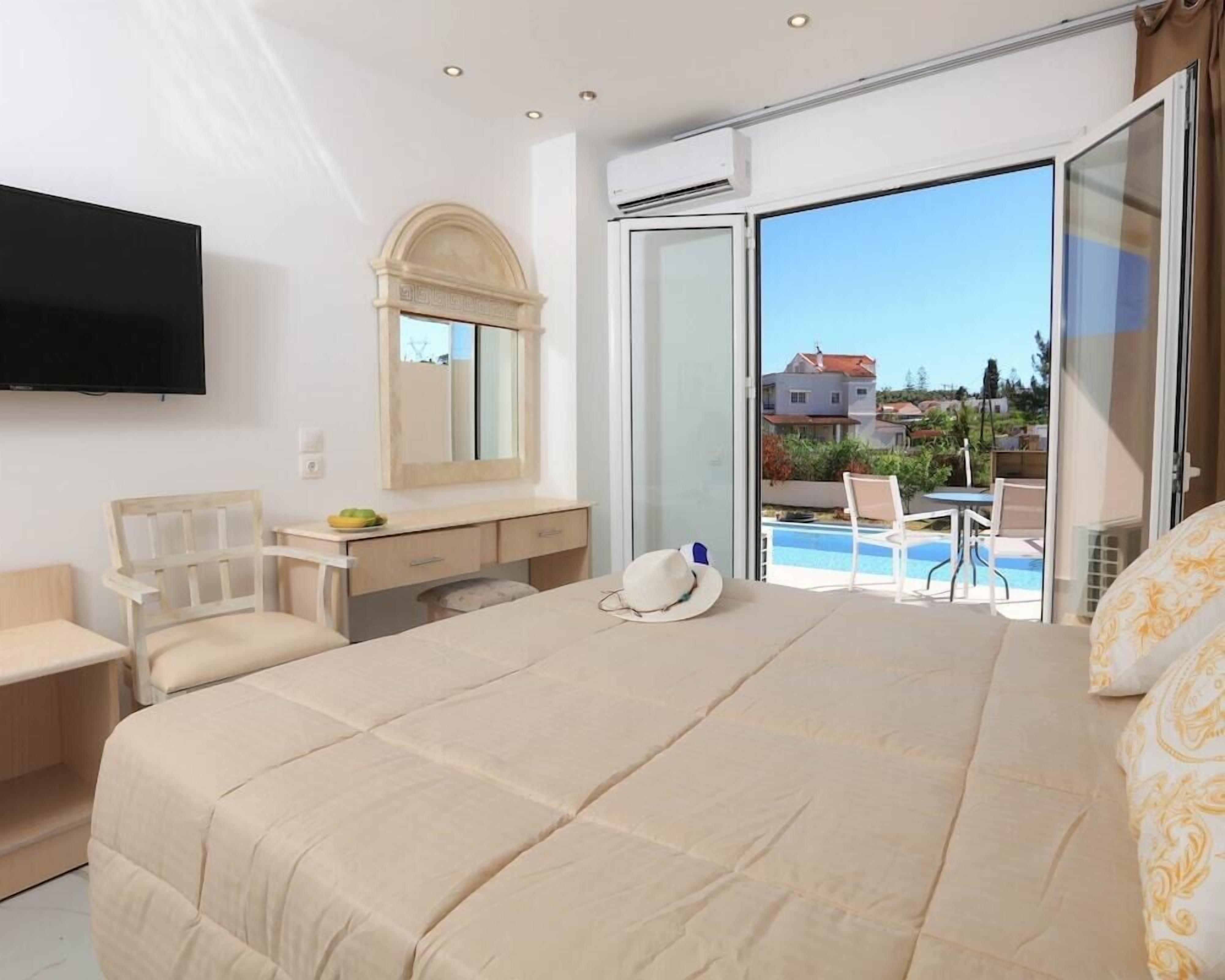 A bedroom with a large bed, a desk, a chair, a TV, and a view of a swimming pool and houses.