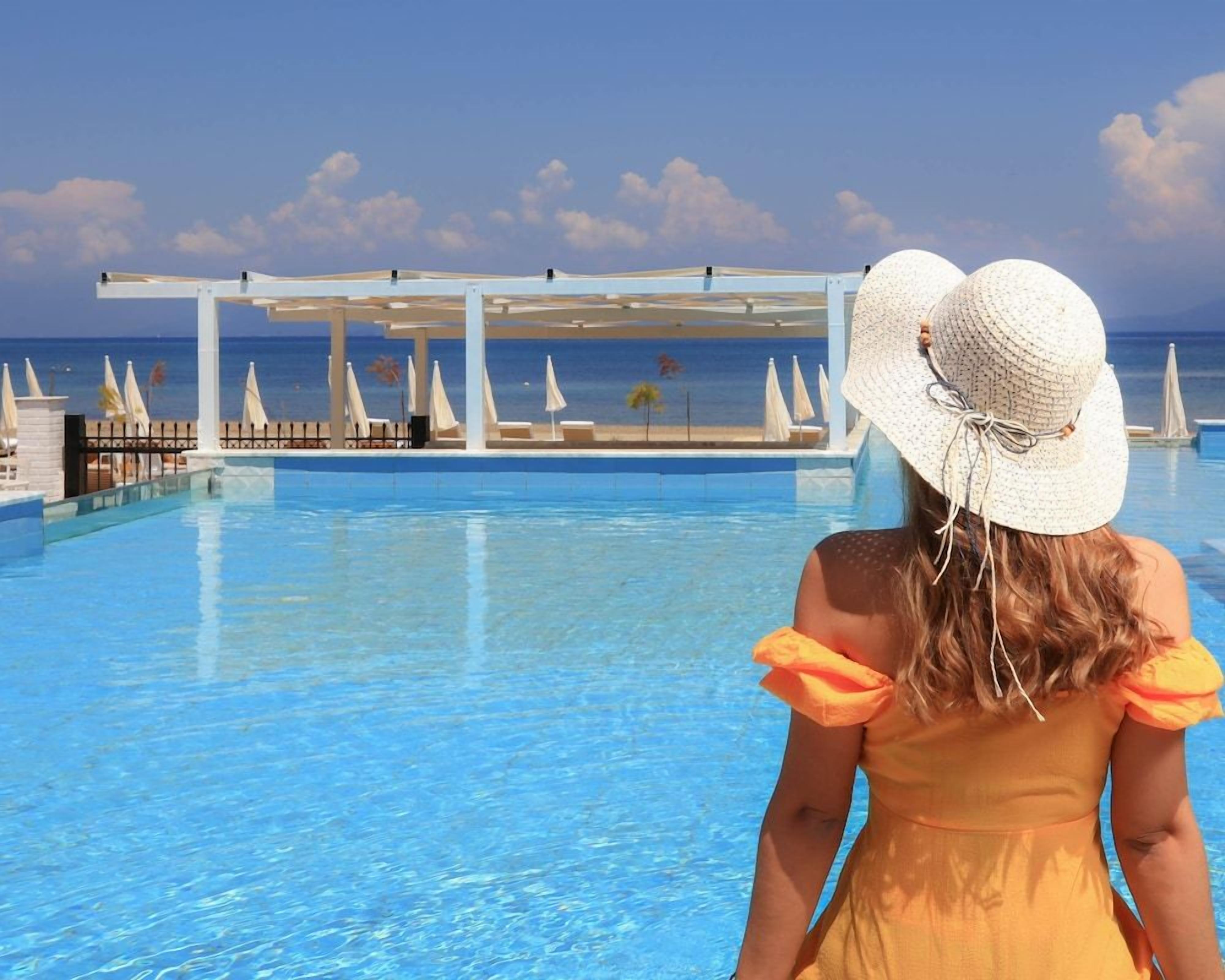A poolside view with a clear sky, a person in a yellow dress and white hat, and a pool with umbrellas and lounge chairs.