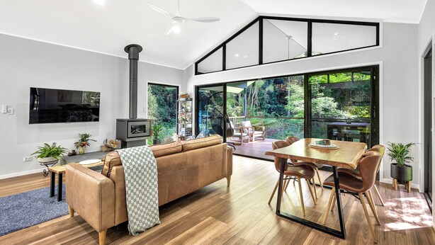 A modern living room with a large sofa, dining table, and chairs. There is a TV mounted on the wall, a fireplace, and large windows offering a view of the outdoors.