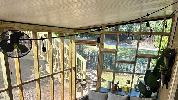 Ein helles Zimmer mit großem Fenster, Korbmöbeln, einem Betontisch und einem kleinen Beistelltisch mit Pflanze.