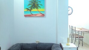 Ein Wohnzimmer mit einer Couch, einem Bild einer Strandansicht, einem Esstisch mit Stühlen und einer Wanduhr.