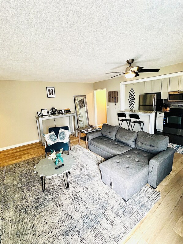 A living room with a grey sectional sofa, a coffee table, and a dining area with a small table and chairs. There is a kitchen area with a microwave, refrigerator, and stove.