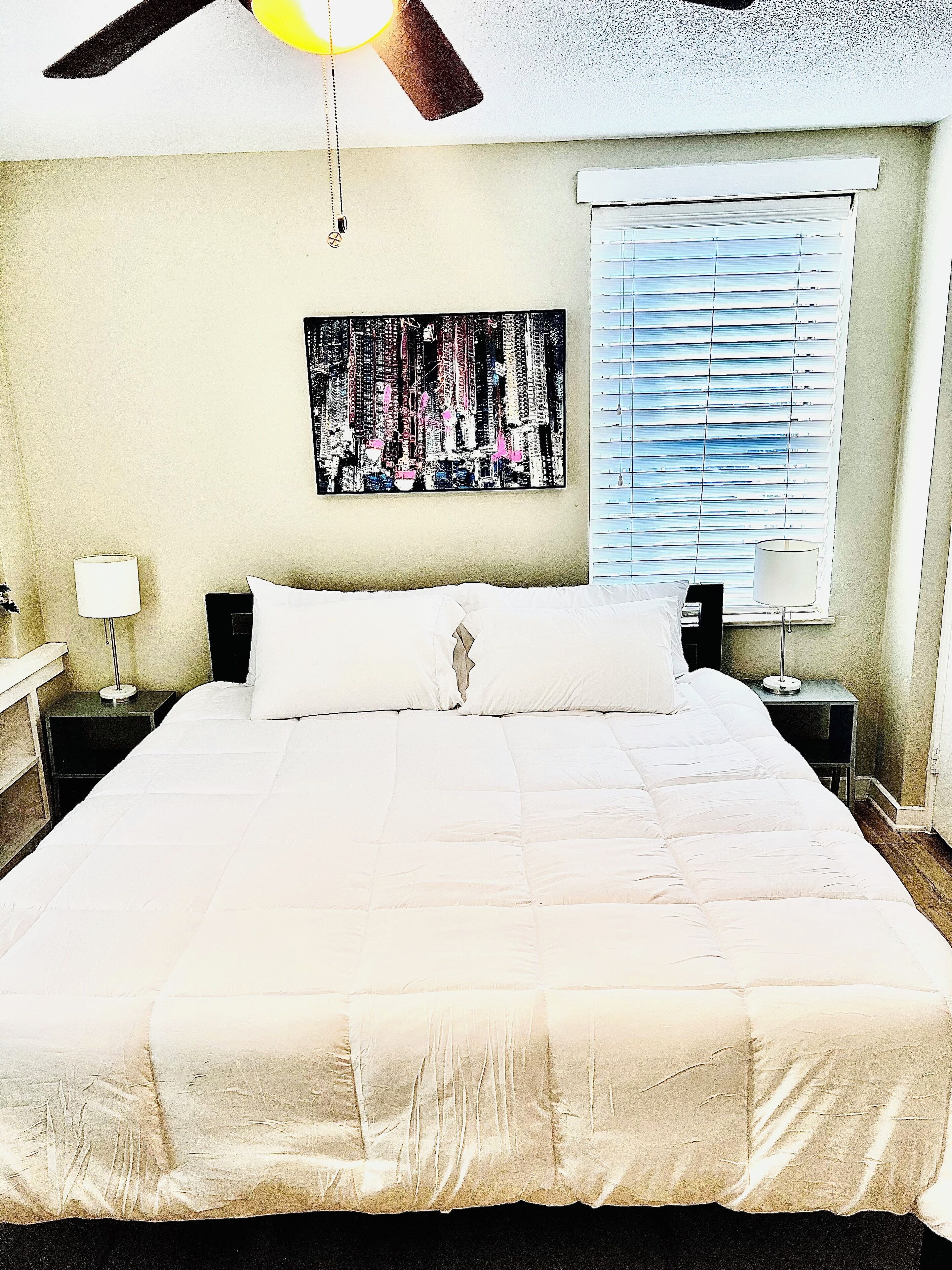 A neatly made bed with white bedding in a room with a ceiling fan, a window with blinds, and a wall-mounted painting.