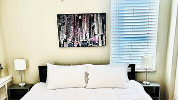 A neatly made bed with white bedding in a room with a ceiling fan, a window with blinds, and a wall-mounted painting.