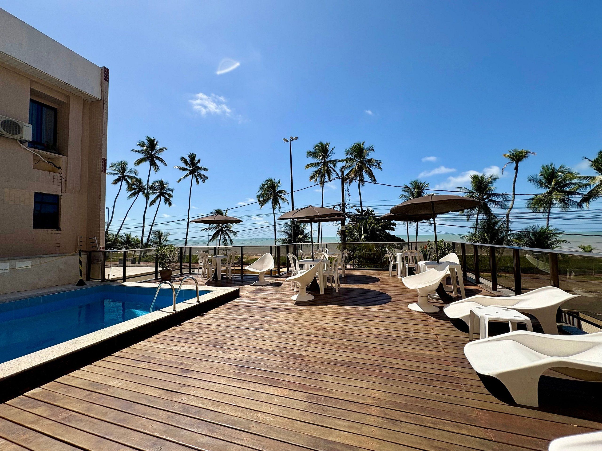 A wooden deck with a pool, white lounge chairs, and umbrellas, overlooking a beach with palm trees.