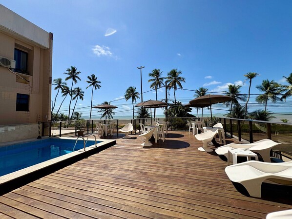 A wooden deck with a pool, white lounge chairs, and umbrellas, overlooking a beach with palm trees. - Boulevard Beira mar do Cabo Branco (João Pessoa)