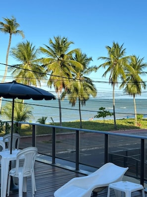A balcony with white plastic chairs and a table, overlooking a beach with palm trees and the ocean. - Boulevard Beira mar do Cabo Branco (João Pessoa)