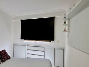 A bedroom with a bed, a TV mounted on the wall, a shelf with decorative items, and a window with a blind. - Boulevard Beira mar do Cabo Branco (João Pessoa)