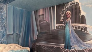 Un dormitori amb llit, un mural amb un personatge i una finestra amb cortines.