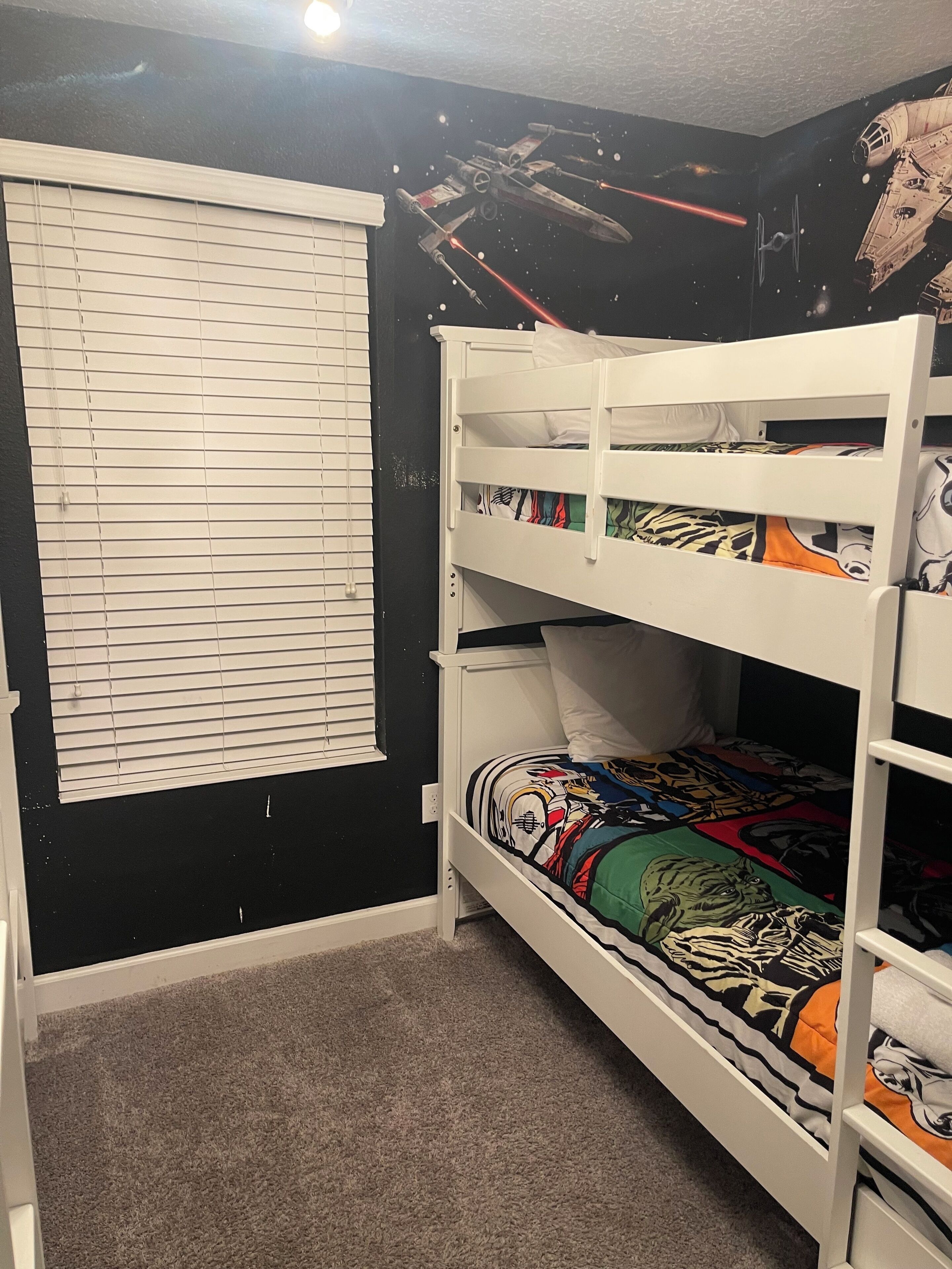 Une chambre avec des lits superposés, un mur décoré d’une fresque sur le thème de Star Wars, une fenêtre avec des stores et un sol recouvert de moquette.