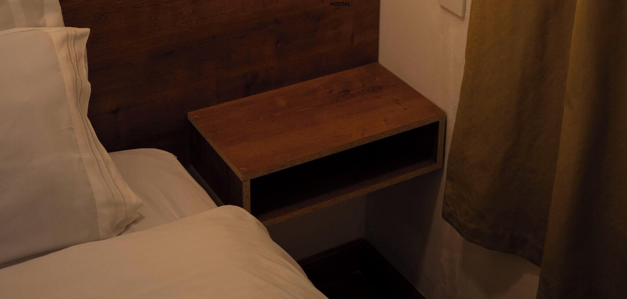 A small, dimly lit room with a wooden headboard, a single bed with white bedding, and a wooden nightstand with a shelf.