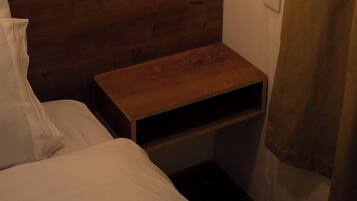 Una habitación pequeña, con poca luz, que presenta un cabecero de madera, una cama individual con ropa blanca y una mesita de noche de madera con un estante.