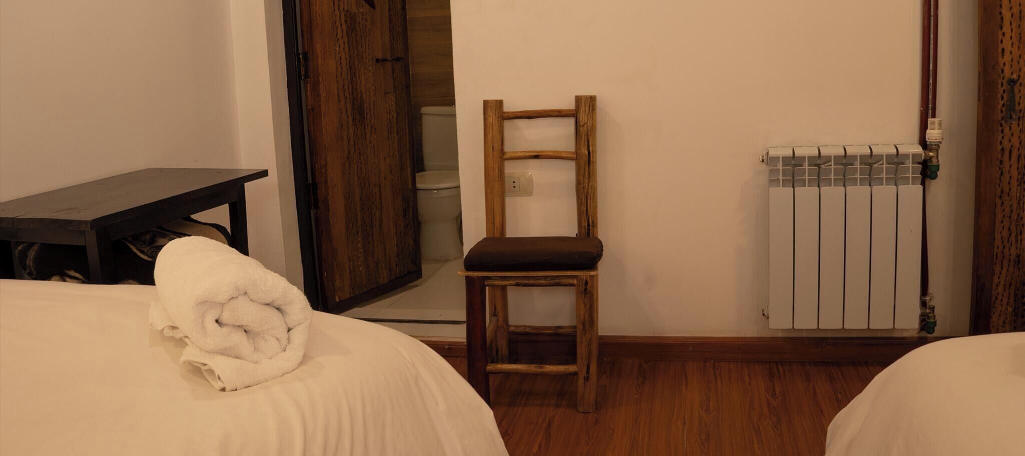 A bedroom with two beds, a wooden chair, a radiator, and a table with a towel.