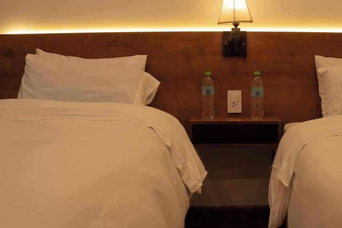 A hotel room with two beds, a nightstand with two water bottles, and a wall-mounted lamp.
