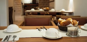 Une table à manger dressée avec des assiettes blanches, des bols et une théière, avec du pain et un panier en arrière-plan.