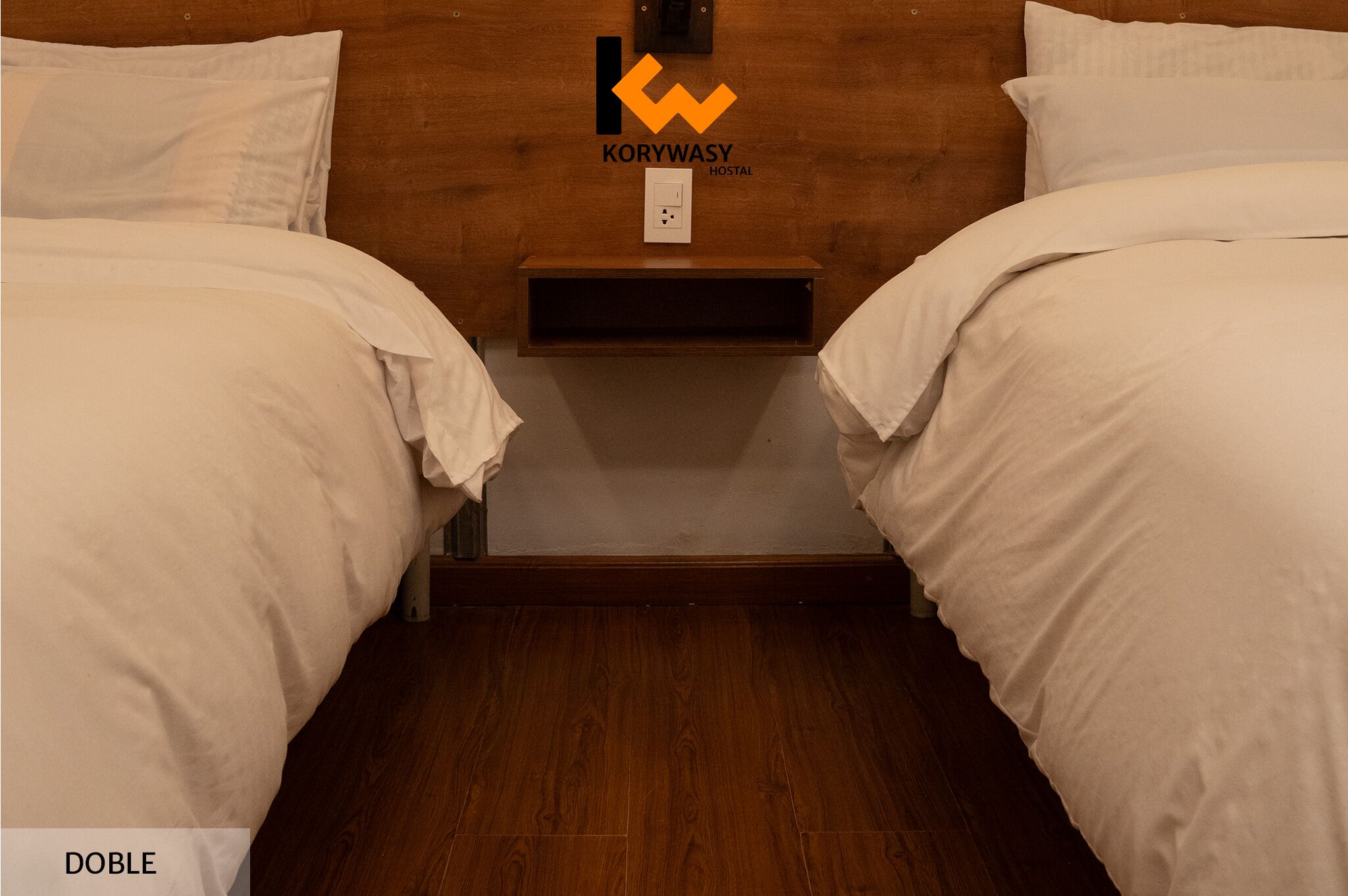 Ein Doppelzimmer mit Holzkopfstütze, weißer Bettwäsche und einem kleinen Regal mit einem Logo.
