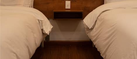 Habitación doble con cabecera de madera, ropa de cama blanca y una pequeña estantería con un logotipo.
